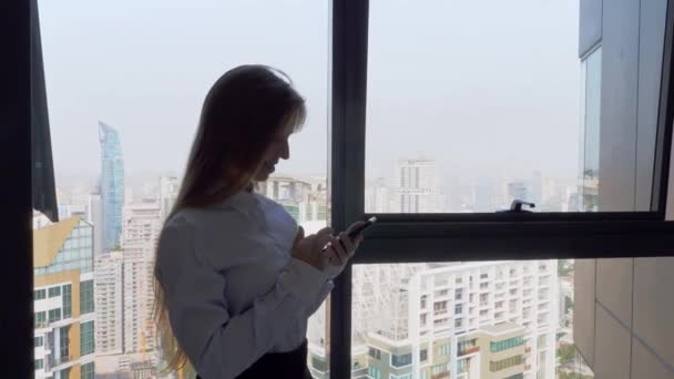 Empresaria de pie en la oficina, trabajando con el teléfono cerca de la ventana con vista a la ciudad — Vídeos de Stock