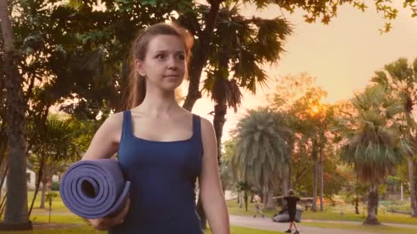Молодая фитнес-женщина в спортивной одежде прогулка в тропическом парке города с ковриком для йоги — стоковое видео