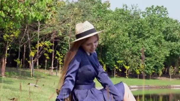 Mujer Turista Sonriente en Vestido y Sombrero de Paja Sentada en el Césped Cerca del Lago — Vídeos de Stock