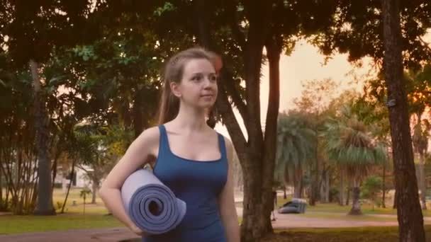 Giovane donna di idoneità in abbigliamento sportivo che cammina nel parco tropicale della città con la stuoia di yoga — Video Stock