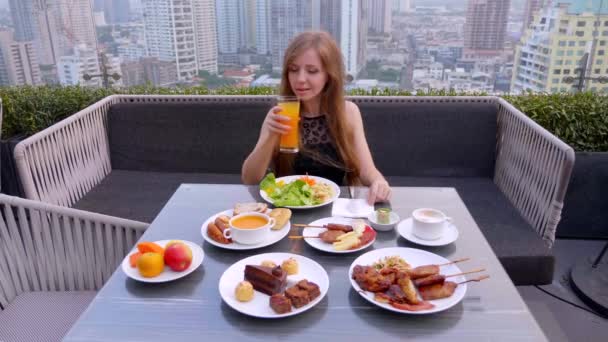 Hermosa chica sentada sola, cenando, bebe jugo de naranja en un bar de la ciudad en la azotea — Vídeos de Stock