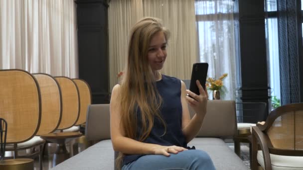 Jovem bonita feliz sentada no sofá e fazendo chamada de vídeo no telefone — Vídeo de Stock