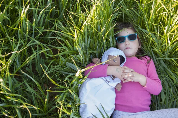 Niña y su muñeca tumbadas en el campo de cereales verde al atardecer — Foto de Stock