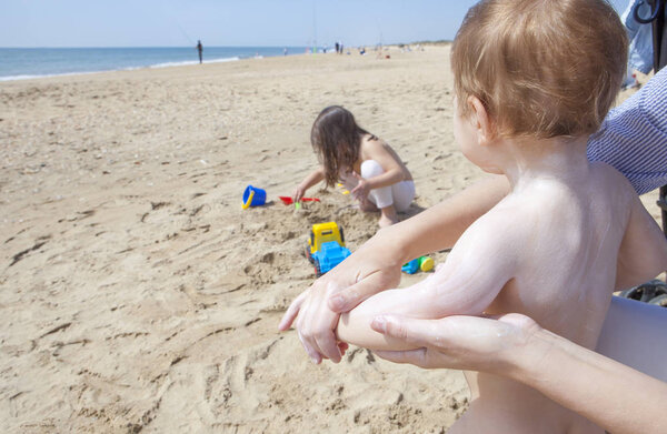 Mother applying sun cream to her baby while her daughter play on