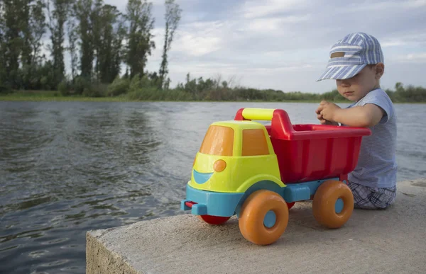 Pequeño niño jugando a la orilla del río con camión volquete cargado con ston — Foto de Stock