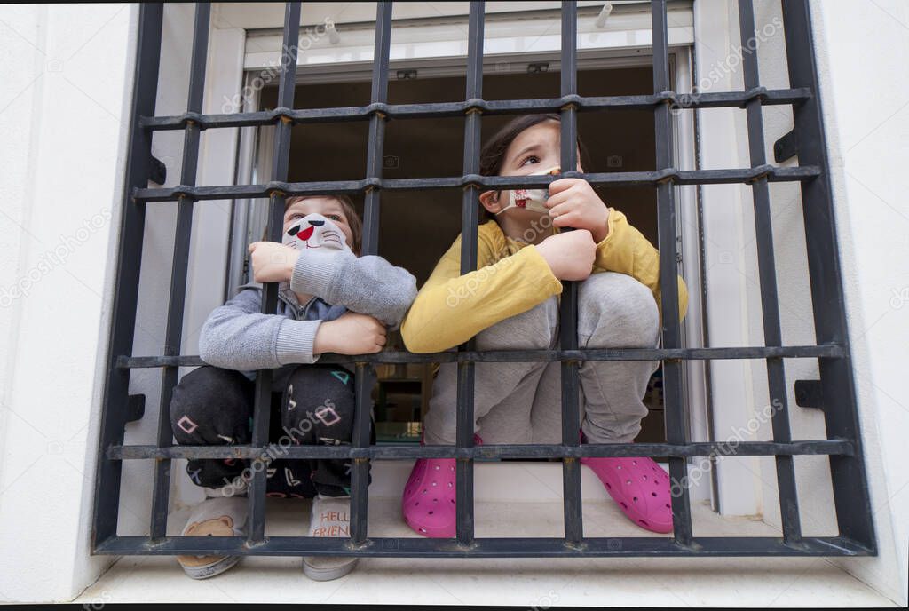 Two children look sadly outdoors through wrought-iron window. Impact on children of Covid-19 confinement
