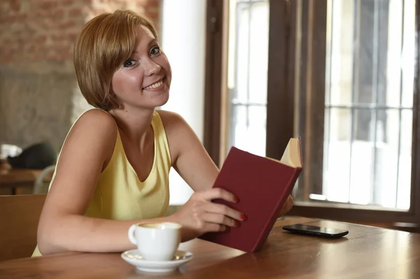 Mujer bonita disfrutando leyendo libro en la cafetería bebiendo taza de café o té sonriendo feliz —  Fotos de Stock