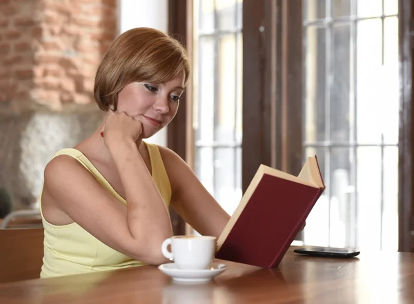 Mooie vrouw geniet van het lezen van boek bij koffiehuis drinken kopje koffie of thee glimlachend gelukkig — Stockfoto