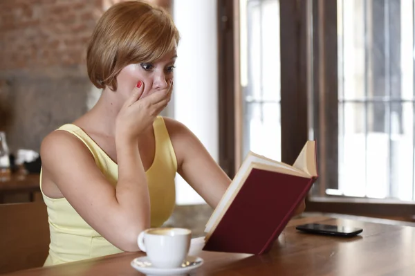 Kaunis nainen nauttii lukeminen kirja kahvilassa juominen kuppi kahvia tai teetä hymyillen onnellinen — kuvapankkivalokuva