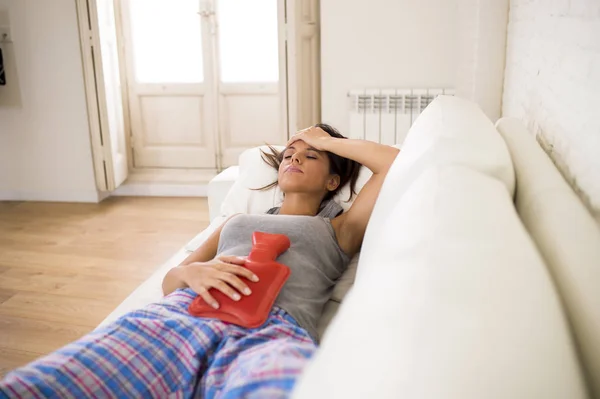 Young beautiful hispanic woman holding hot water bottle against belly suffering menstrual period pain — Stock Photo, Image