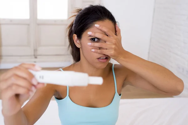 Mulher latina segurando teste de gravidez na cama em casa olhando para o resultado positivo em choque e estresse — Fotografia de Stock