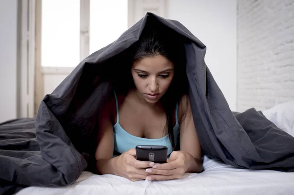 Молодая красивая латиноамериканка, зависимая от мобильного телефона, лежит в постели с одеялом над головой и пишет смс-ки в интернете — стоковое фото