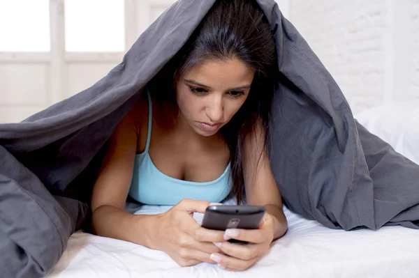 Молодая красивая латиноамериканка, зависимая от мобильного телефона, лежит в постели с одеялом над головой и пишет смс-ки в интернете — стоковое фото
