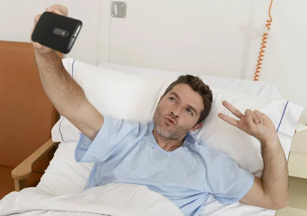 Atraktivní muž ležící na posteli nemocnice klinika držení mobilního telefonu, přičemž autoportrét selfie Foto — Stock fotografie