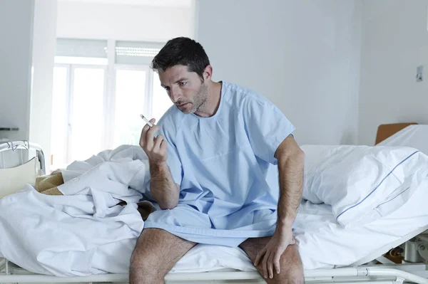 Aantrekkelijke jongeman triest en ongerust kijken naar ziekenhuis bed roken sigaret in kliniek slaapkamer — Stockfoto