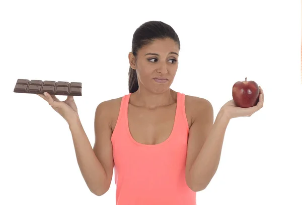 Young attractive hispanic woman in fitness top holding apple fruit and chocolate bar in her hands — Stock Photo, Image