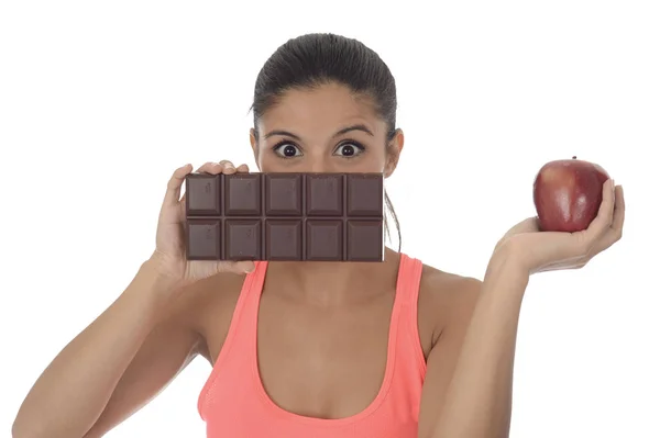 Young attractive hispanic woman in fitness top holding apple fruit and chocolate bar in her hands — Stock Photo, Image