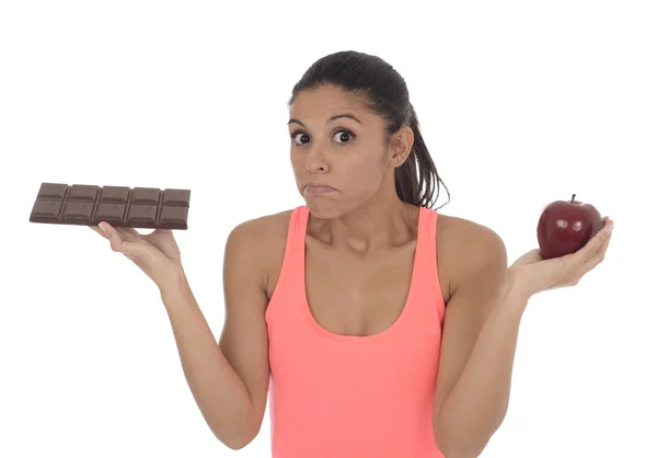 Jovem mulher hispânica atraente em fitness top segurando fruta de maçã e barra de chocolate em suas mãos — Fotografia de Stock