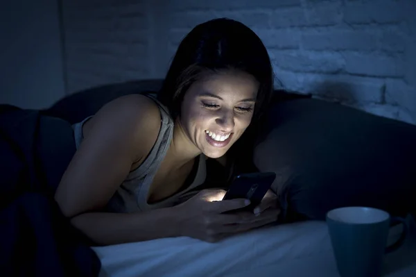 Chica en la cama usando el teléfono móvil tarde en la noche en dormitorio oscuro acostado feliz y relajado —  Fotos de Stock