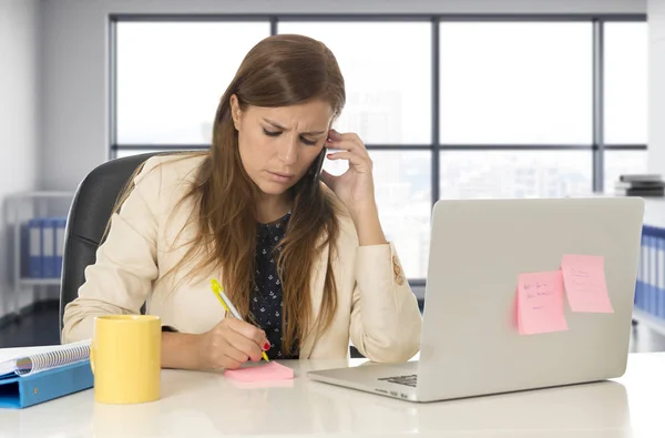 Напряженная женщина, работающая с ноутбуком на столе, разговаривает на мобильном телефоне, записывает — стоковое фото