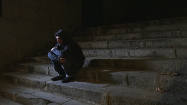 4K video lateral panning 24 fps of young desperate wasted man in hood suffering stress and drepression sitting suffer on urban street staircase at night sad and worry in addiction concept — Stok Video