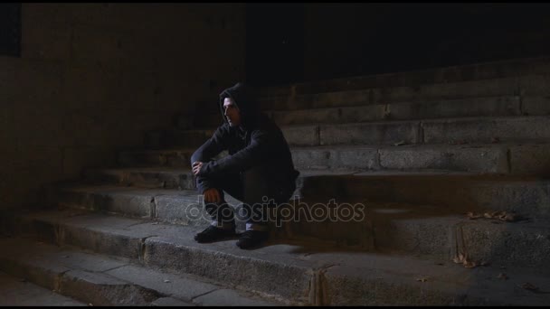 4K video lateral panning 24 fps of young desperate wasted man in hood suffering stress and drepression sitting miserable on urban street staircase at night sad and worried in addiction concept — Stock Video