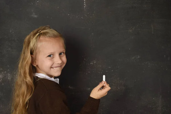 Hermosa rubia dulce colegiala en uniforme celebración tiza escritura en pizarra sonriendo feliz — Foto de Stock