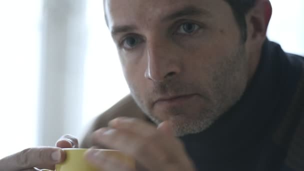 4k 24 fps Handkamera-Tracking auf junge traurige unrasierte Mann zu Hause sitzt vor dem Fenster und sieht traurig und deprimiert trinken Tasse Kaffee leiden Depressionen Konzept und Probleme — Stockvideo