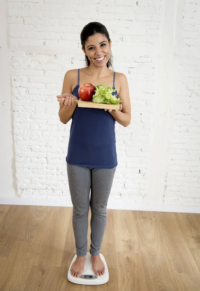 Attractive slim latin woman or teenager girl checking weight on scale holding vegetable tray — Stock Photo, Image