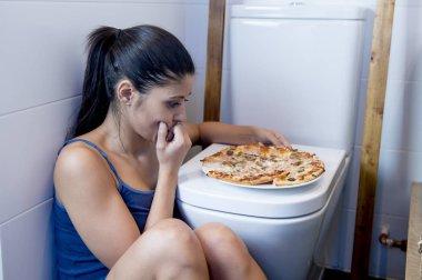bulimic woman feeling sick guilty sitting at the floor of the toilet leaning on WC eating pizza clipart