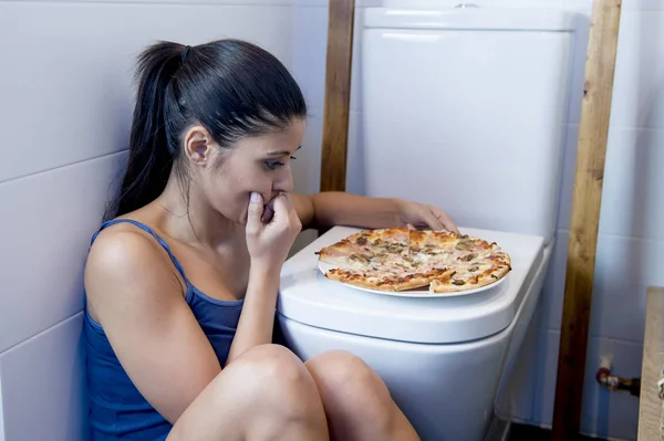 Булимия женщина чувствует себя виноватой, сидя на полу унитаза опираясь на туалет есть пиццу — стоковое фото