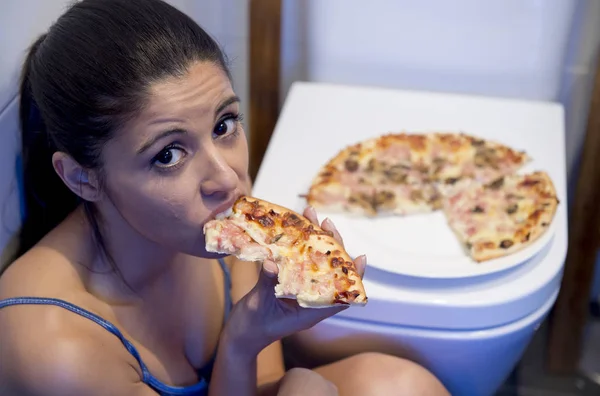 Bulimie-Frau, die sich schuldig fühlt, sitzt auf dem Boden der Toilette und lehnt sich an die Toilette und isst Pizza — Stockfoto