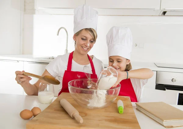 Young mother and little sweet daughter in cook hat and apron cooking together baking at home kitchen — Stock Photo, Image