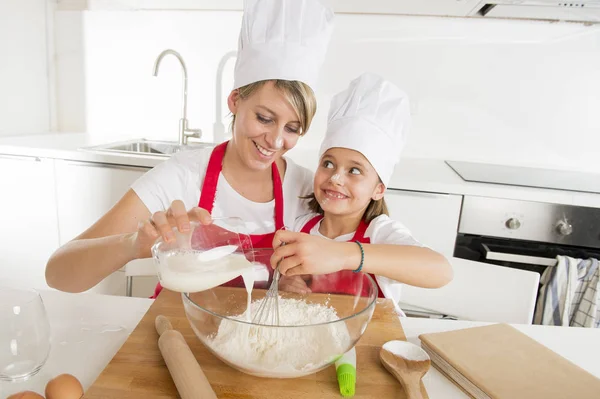 Young mother and little sweet daughter in cook hat and apron cooking together baking at home kitchen — Stock Photo, Image