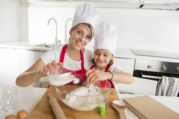 Young mother and little sweet daughter in cook hat and apron cooking together baking at home kitchen — Stock Photo, Image