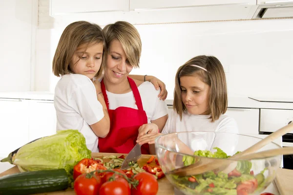 Joven madre en delantal de cocinero y dulces hermosas hijas gemelas cocinando preparando juntos ensalada — Foto de Stock