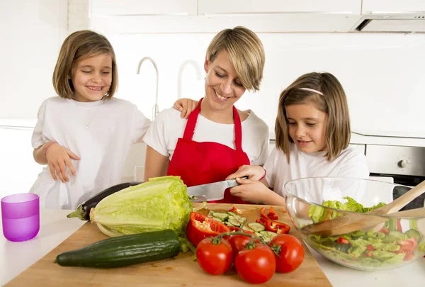 Jonge moeder in cook schort en zoete mooie tweelingdochters koken voorbereiding samen salade — Stockfoto
