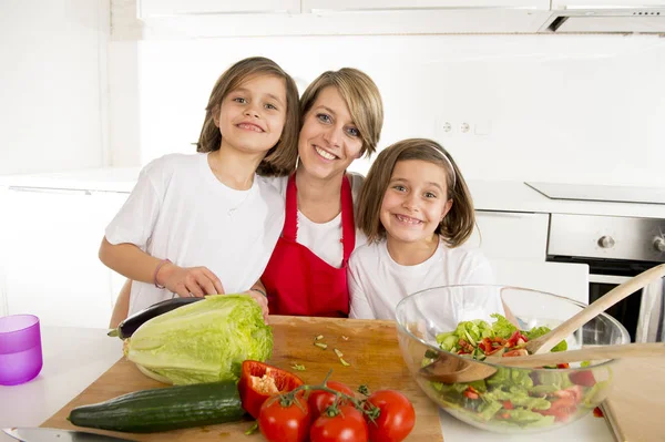 Joven madre en delantal de cocinero y dulces hermosas hijas gemelas cocinando preparando juntos ensalada — Foto de Stock
