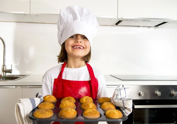 Little and cute girl alone in cook hat and apron presenting and showing tray with muffins smiling happy — Stock Photo, Image