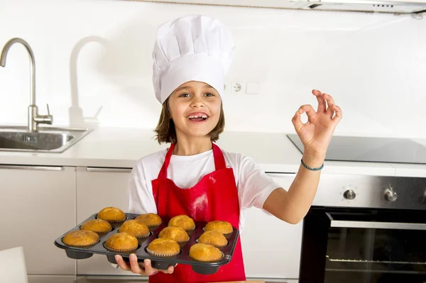 Little and cute girl alone in cook hat and apron presenting and showing tray with muffins smiling happy — Stock Photo, Image