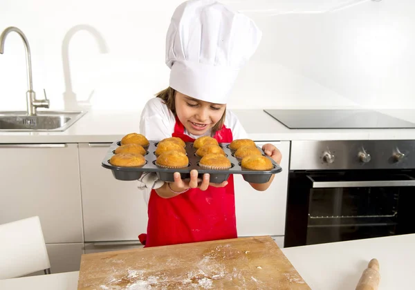 Little and cute girl alone in cook hat and apron presenting and showing tray with muffins smiling happy — Stock Photo, Image