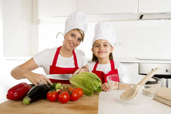 Mãe e pequena filha cozinhar em conjunto com chapéu avental preparando salada em casa cozinha — Fotografia de Stock