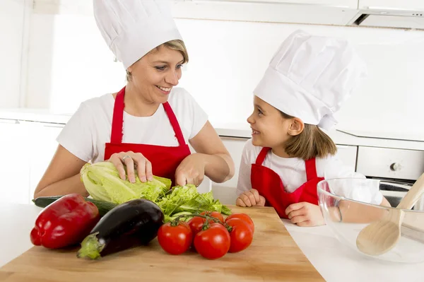 Mother and little daughter cooking together with hat apron preparing salad at home kitchen — Stock Photo, Image