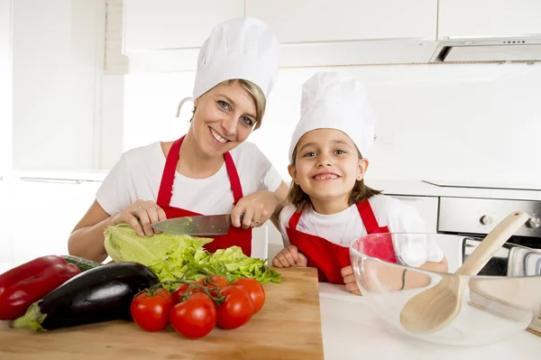 Madre e hija pequeña cocinando junto con delantal de sombrero preparando ensalada en la cocina casera —  Fotos de Stock