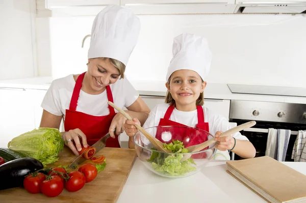 Mother and little daughter cooking together with hat apron preparing salad at home kitchen — Stock Photo, Image