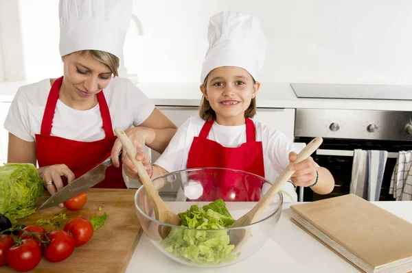 Mãe e pequena filha cozinhar em conjunto com chapéu avental preparando salada em casa cozinha — Fotografia de Stock