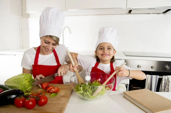 Mother and little daughter cooking together with hat apron preparing salad at home kitchen — Stock Photo, Image
