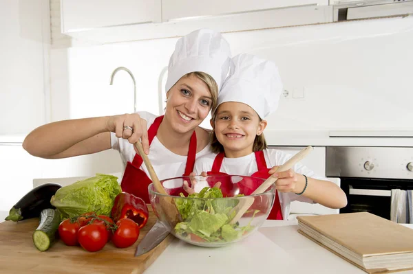 Mãe e pequena filha cozinhar em conjunto com chapéu avental preparando salada em casa cozinha — Fotografia de Stock