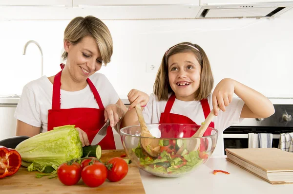 Madre e hija pequeña cocinando junto con delantal cocinero preparando ensalada en la cocina casera —  Fotos de Stock