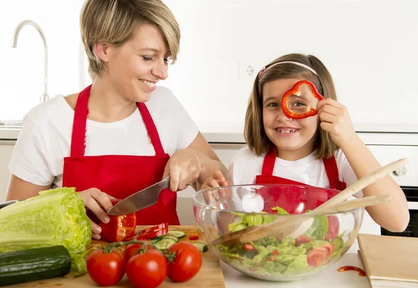 Madre e hija pequeña cocinando junto con delantal cocinero preparando ensalada en la cocina casera —  Fotos de Stock